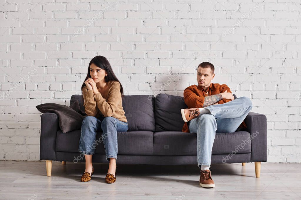 Young man looking at offended asian wife sitting on couch in living room, relationship trouble