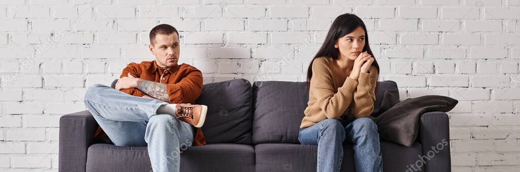 Husband looking at offended asian wife sitting on couch in living room, horizontal banner
