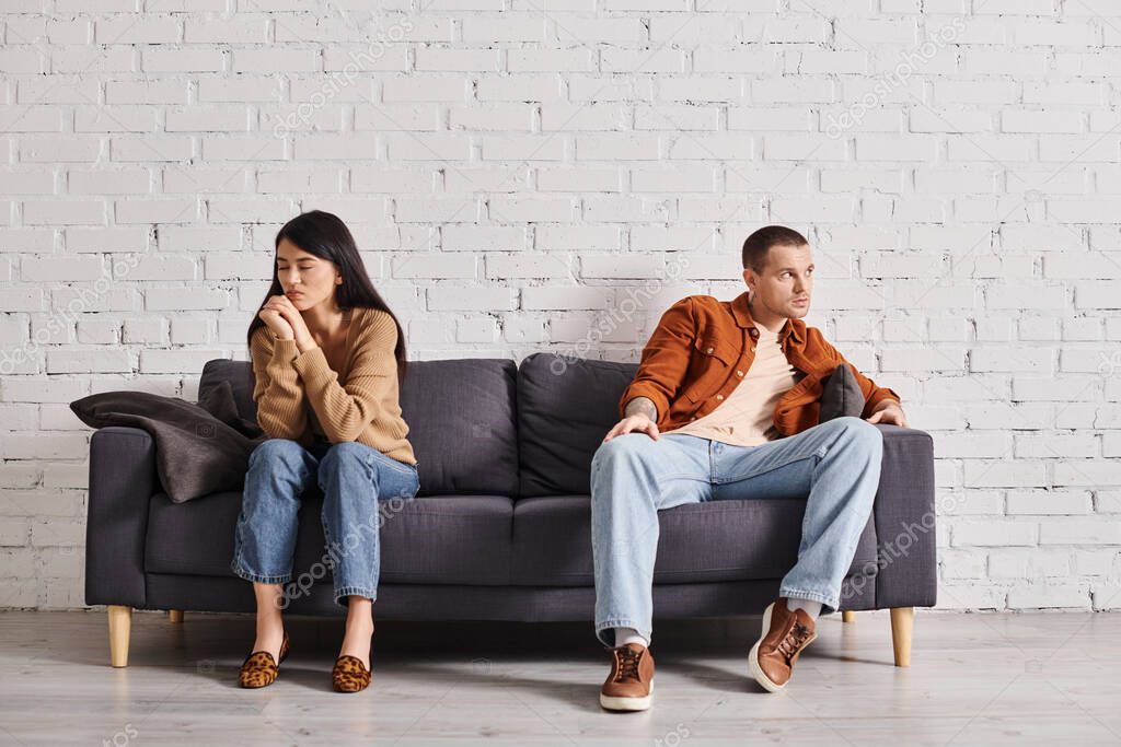 Depressed interracial couple looking away while sitting on couch in living room, divorce concept