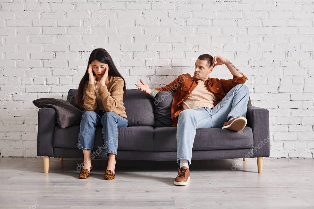 Young man talking to offended asian woman on couch in living room, relationship difficulties