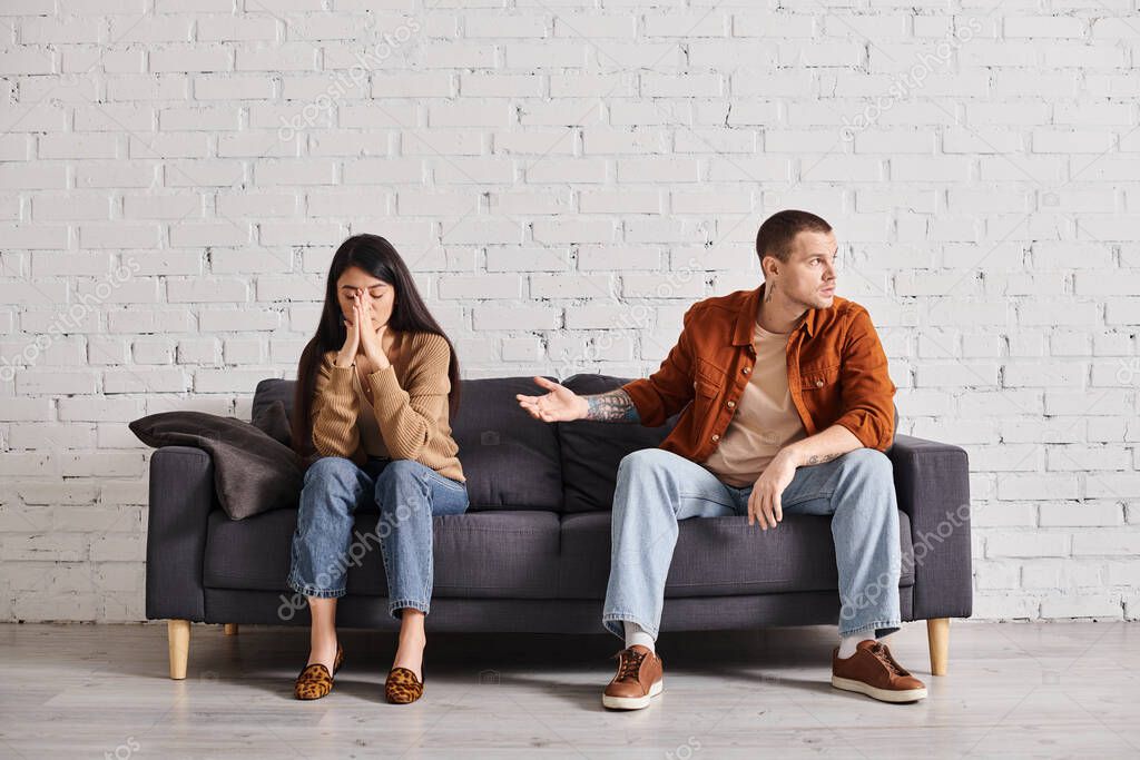 Discouraged man pointing at offended asian wife on couch in living room, relationship difficulties