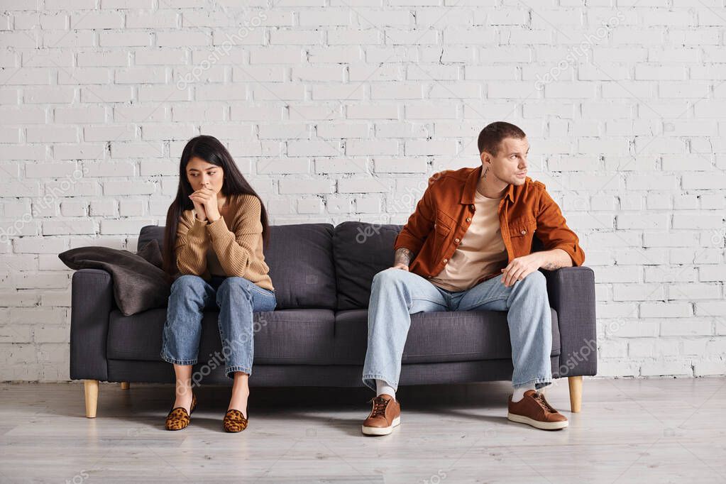 Young interracial offended couple sitting on couch in living room, heartache misunderstanding