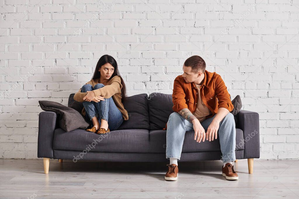Frustrated interracial couple looking at each other on couch in living room, divorce concept
