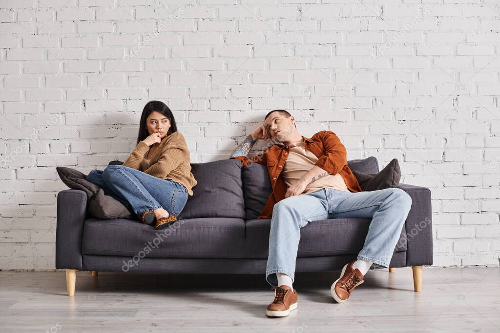 Young self-confident man talking to upset asian wife on couch in living room, divorce concept