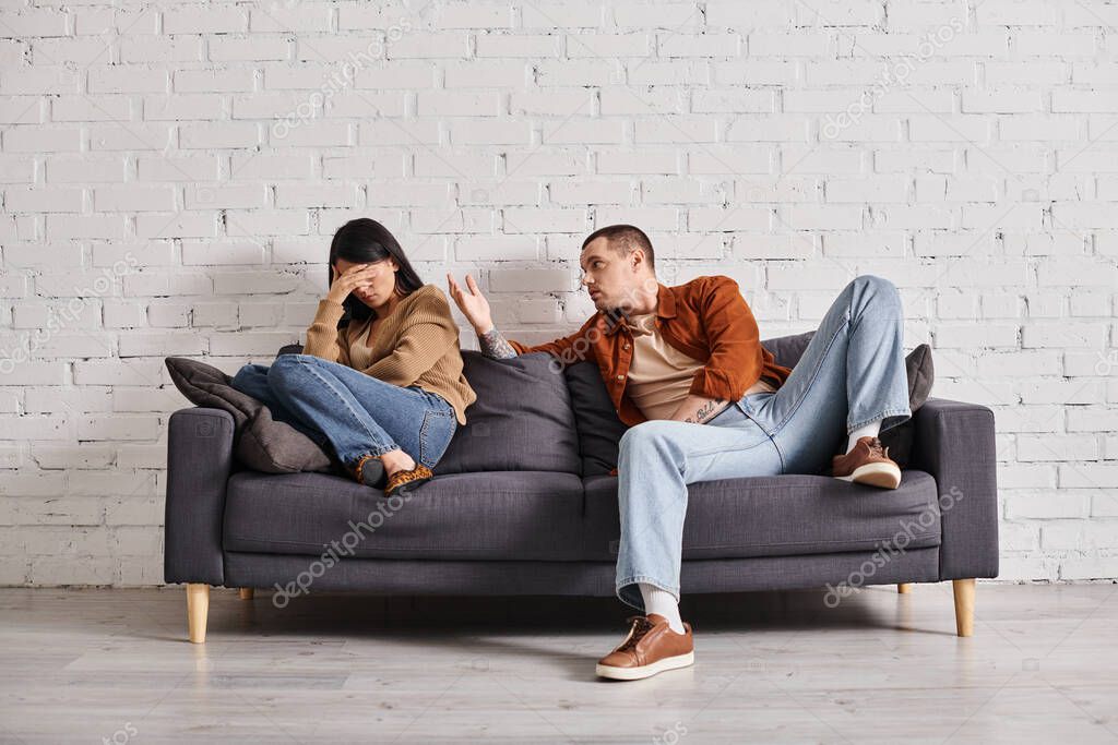 Young emotional man talking to asian woman crying on couch in living room, divorce concept