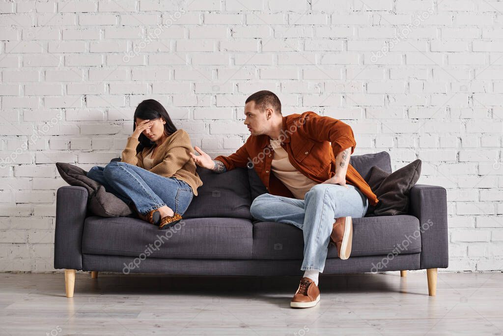 Young irritated man talking to young asian wife crying on couch in living room, divorce concept