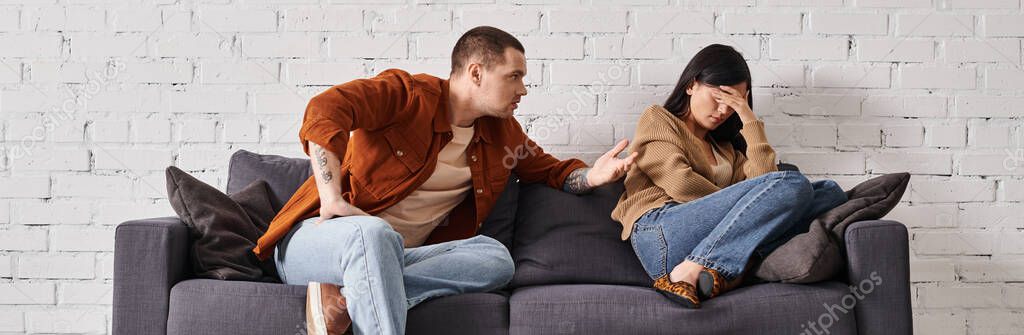 Irritated husband talking to young asian wife crying on couch in living room, horizontal banner