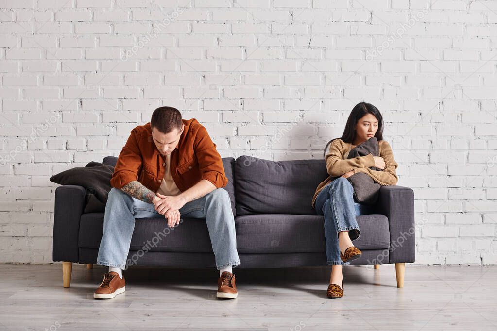 Young offended interracial couple sitting on couch in modern living room, family divorce concept