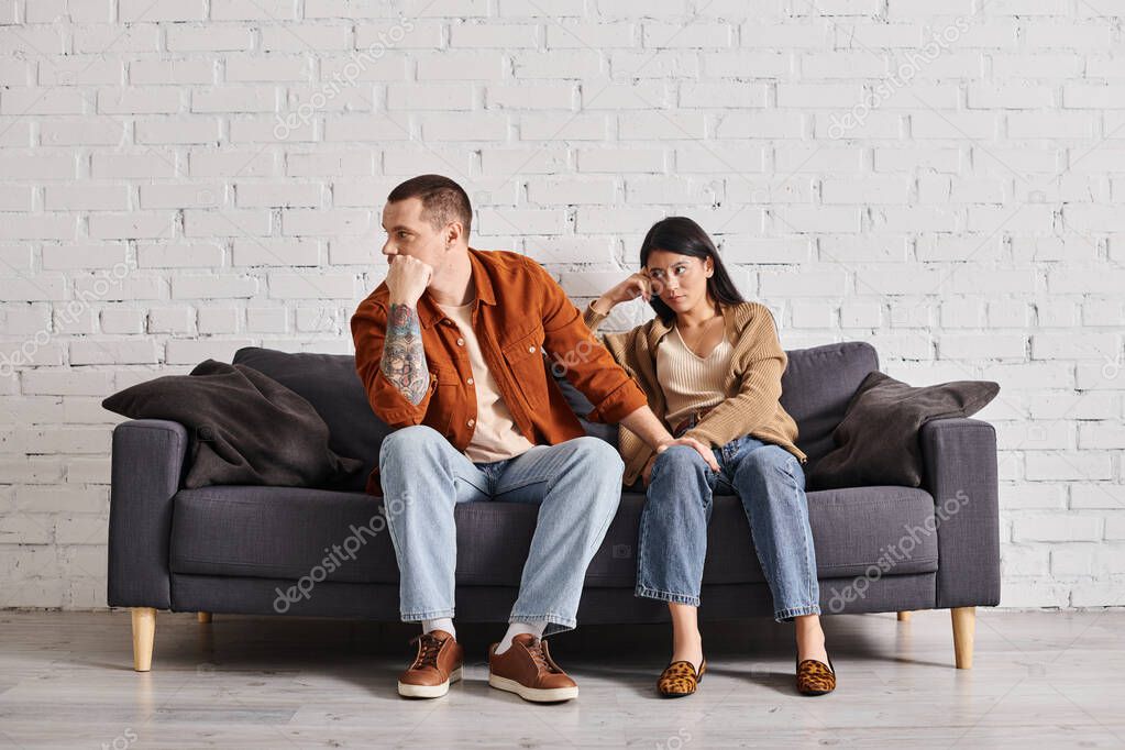 Young interracial couple in quarrel sitting on couch in living room at home, divorce concept