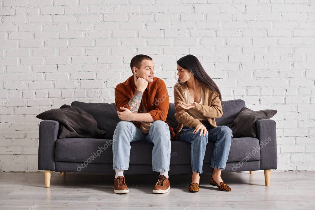 Family divorce, offended asian woman talking to smiling skeptical husband on couch in living room