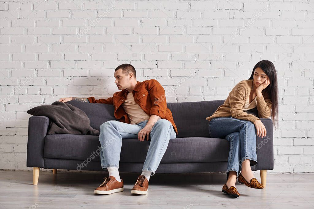 Young interracial couple with emotional distress sitting on couch in living room, divorce concept