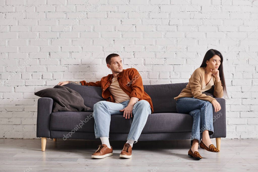 Young man looking at offended asian wife while sitting on couch in living room, divorce concept