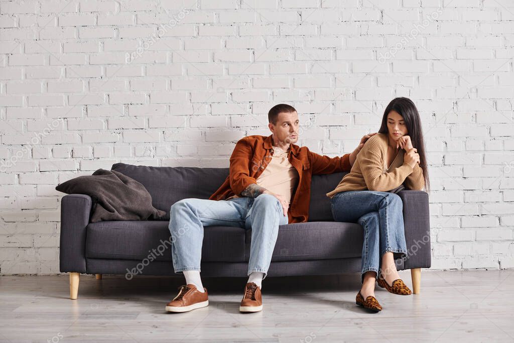 Young man calming upset asian wife sitting on couch in living room, relationship difficulties