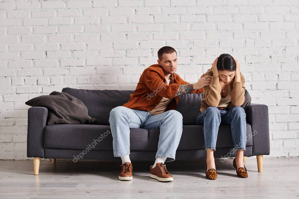 Husband calming frustrated asian wife sitting on couch in living room, relationship difficulties