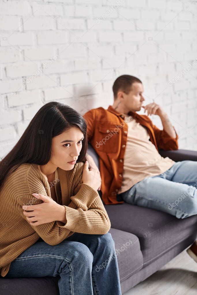 Young frustrated asian woman sitting with folded arms near upset husband on couch in living room