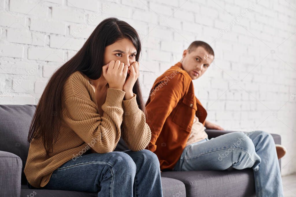 Displeased man looking at offended asian wife on couch in living room, relationship difficulties