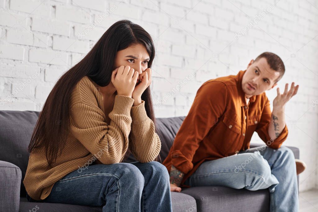 Discouraged man talking to offended asian wife sitting on couch in living room, divorce concept