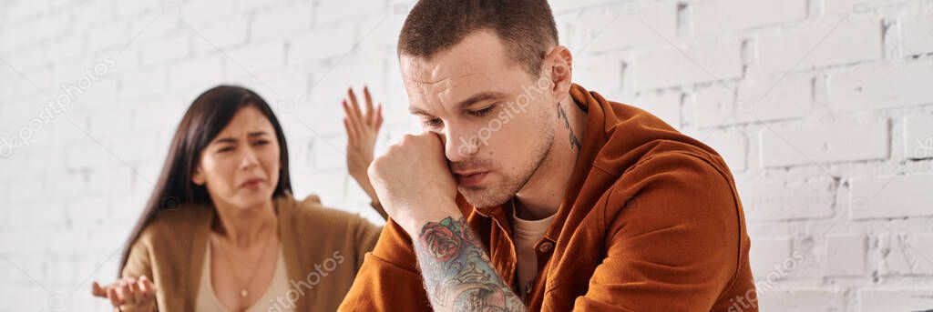 Stressed asian woman quarrelling at offended tattooed husband on couch at home, banner