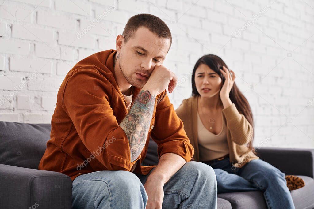 Frustrated asian woman looking at sad tattooed husband on couch at home, family divorce concept