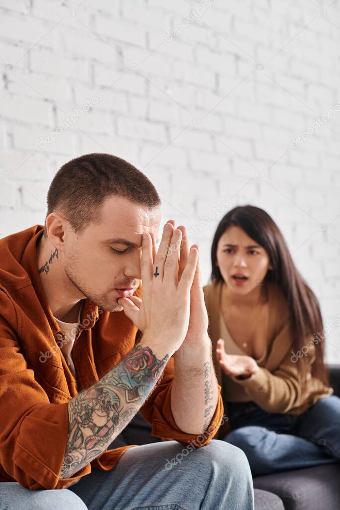 Young tattooed man sitting with closed eyes near angry asian man quarrelling at home, divorce