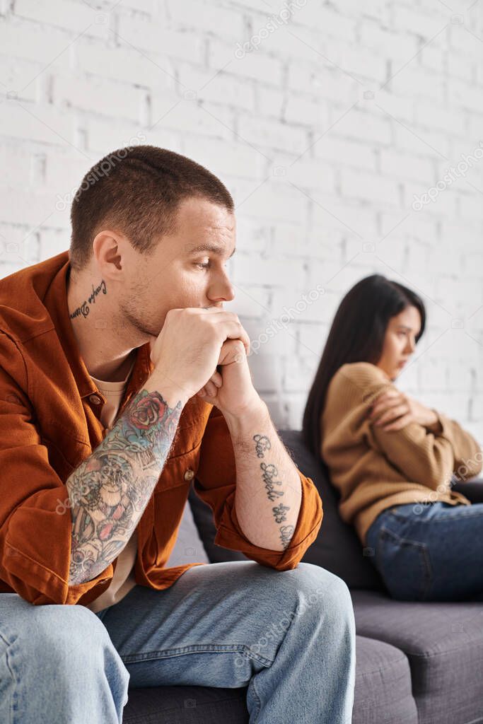 Frustrated tattooed man looking at offended asian wife sitting on couch at home, divorce concept