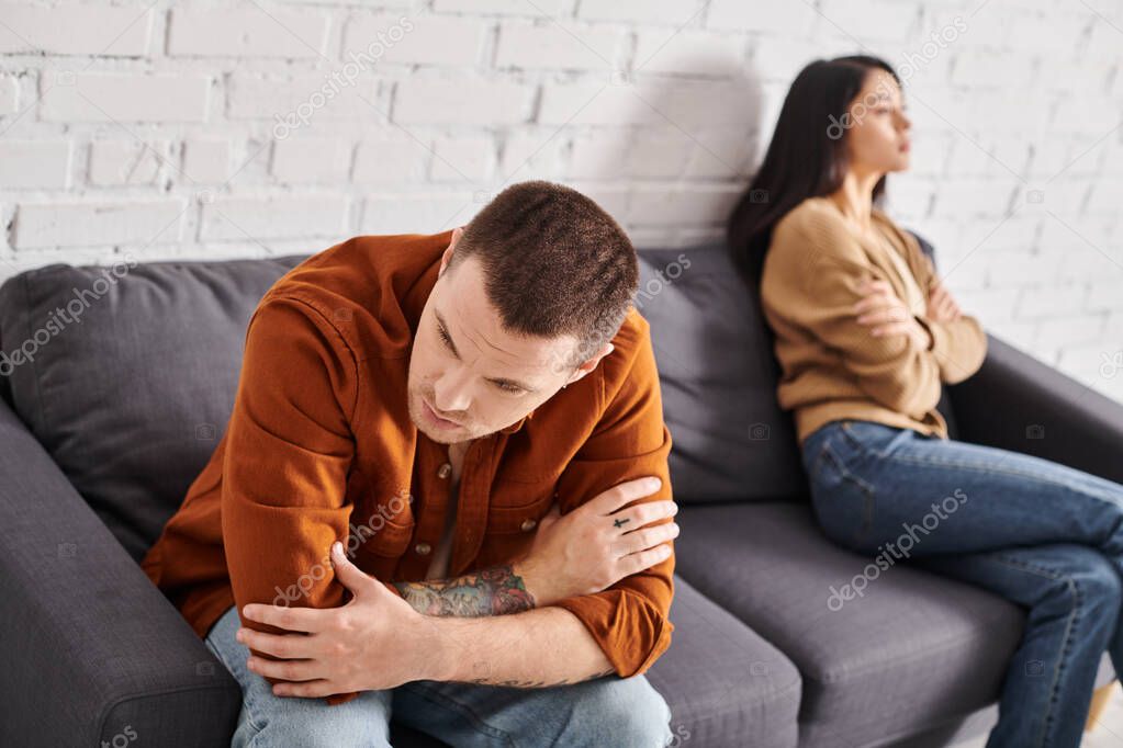 Gloomy tattooed man sitting on couch in living room near offended asian wife, family problem