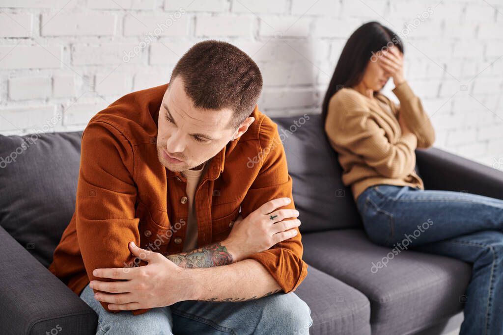 Upset tattooed man sitting on couch in living room near crying asian wife, divorce concept