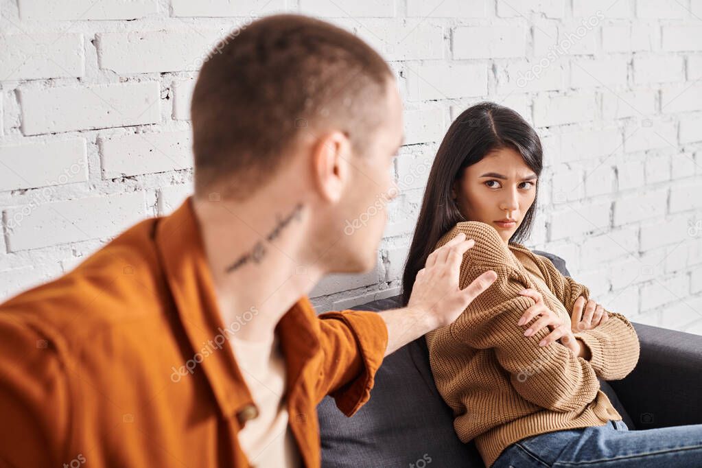 Young man trying to calm offended asian wife sitting on couch at home, relationship trouble