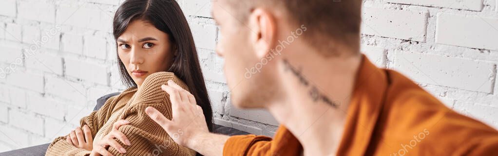 Young tattooed man trying to calm upset asian wife sitting on couch at home, horizontal banner