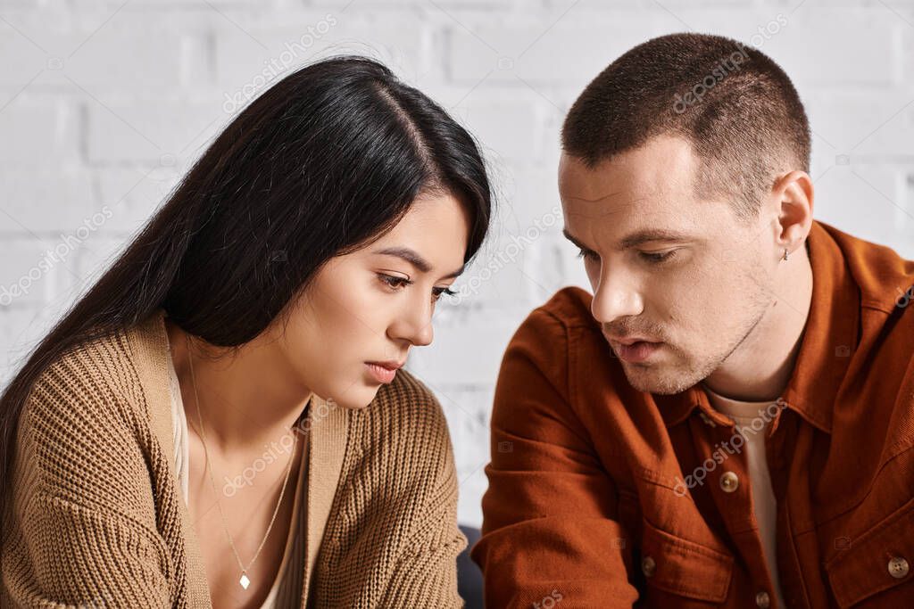 Young and frustrated multiethnic couple talking in living room at home, relationship difficulties