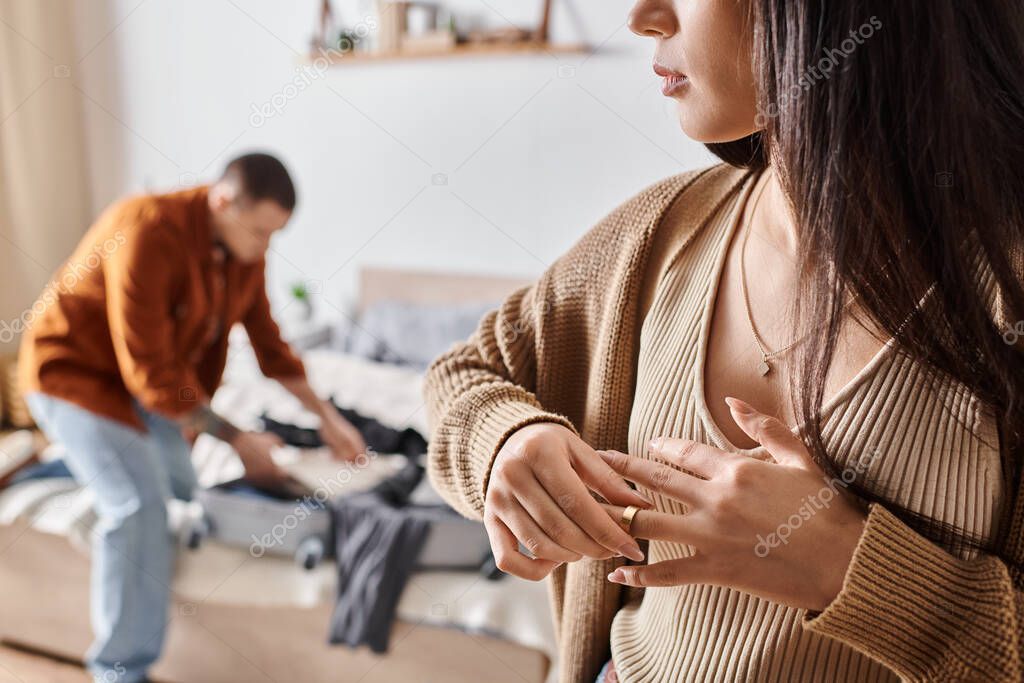 Young woman taking off wedding ring while husband packing suitcase in bedroom, divorce concept