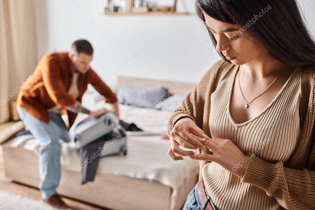 Asian woman taking off wedding ring while husband packing suitcase in bedroom, family divorce