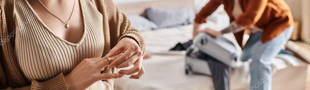Cropped view of young wife taking off wedding ring near husband packing suitcase in bedroom, banner