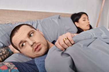 young offended tattooed man looking away while lying down on bed near asian wife, family trouble clipart