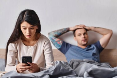 Şaşırmış Asyalı bir kadın kocasının yanındaki akıllı telefona bakıyor evde yatakta yatıyor, aile içi çatışmalar var.