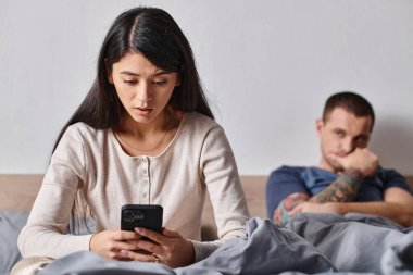 Şok olmuş Asyalı kadın kocasının yanındaki akıllı telefona bakıyor evde yatakta yatıyor, aile içi çatışmalar var.