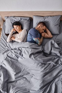 high angle view of depressed interracial couple lying down on bed at home, family divorce concept clipart