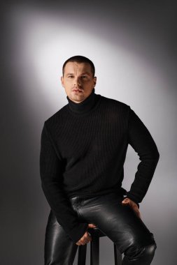 young fashionable man in black turtleneck sitting and looking at camera on grey with lighting clipart