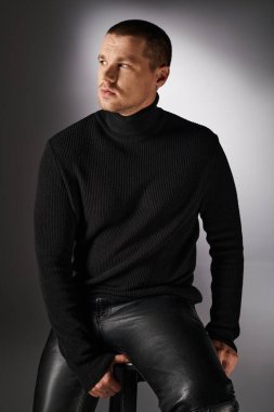 pensive trendy man in black turtleneck sitting and looking away on grey backdrop with lighting clipart