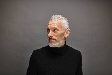 İyi giyimli, çekici, balıkçı yaka kazaklı, gri arka planda poz veren ve başka yöne bakan olgun bir adam.