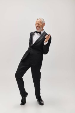good looking joyous mature man with gray beard and bow tie in elegant tuxedo dancing actively clipart