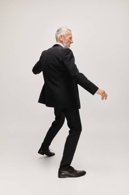 Şık takım elbiseli neşeli, yakışıklı, olgun bir adam papyonlu, gri arka planda mutlu bir şekilde dans ediyor.