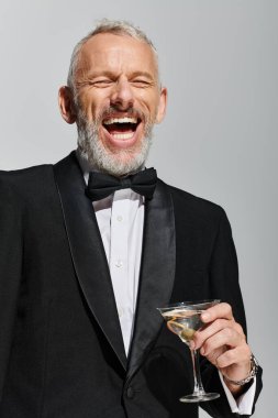 Mutlu, olgun, sakallı, zarif smokinli, elinde martini olan ve neşeyle gülümseyen bir adam.