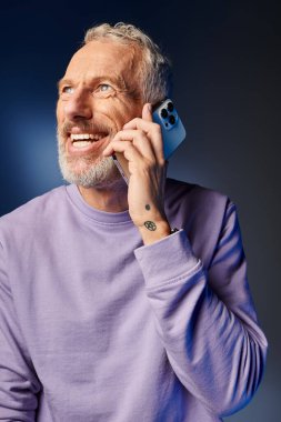 Neşeli sakallı, modaya uygun, mor renkli, şık kazaklı olgun bir adam mavi arka planda telefonla konuşuyor.