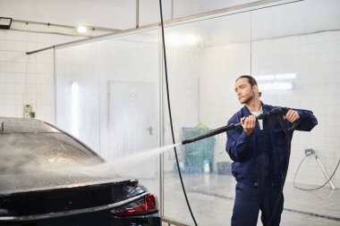 appealing devoted serviceman in comfy uniform with collected hair washing modern car in garage clipart