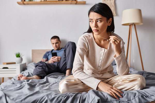 upset asian woman sitting near husband using smartphone in bedroom at home, family problem