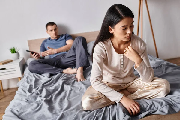 stock image frustrated asian woman sitting near husband using smartphone in bedroom at home, family problem