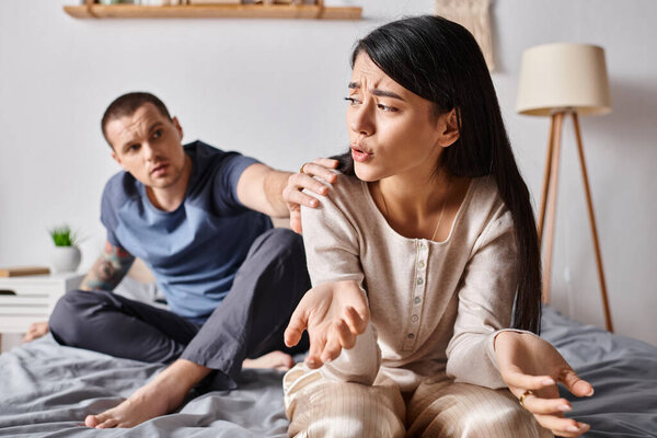 молодой татуированный мужчина разговаривает с расстроенной азиатской женой в спальне дома, трудности в отношениях