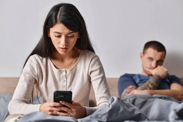 stock image shocked asian woman looking at smartphone near husband lying on bed at home, family conflict