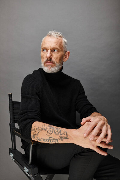 good looking bearded mature man in fashionable turtleneck sitting on folding chair and looking away